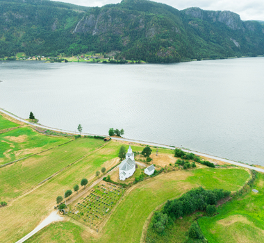 Aerial view of the white church and its cemetery situated in green fields beside a large lake with mountains in the background in Araksbo Agder Norway.