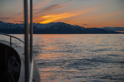 View of the serene water reflecting the fiery sunset sky with snow capped mountains in the background from a boat H&uacute;sav&iacute;k Nor&eth;ur&thorn;ing Iceland.