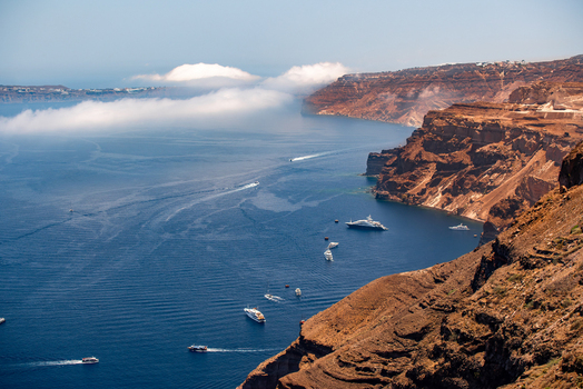 View of the dramatic volcanic cliffs of Fira overlooking the deep blue Aegean Sea with several yachts and boats sailing near the coastline in Fira Greece. View of the dramatic volcanic cliffs of Fira overlooking the deep blue Aegean Sea with several yachts and boats sailing near the coastline in Fira Greece.
