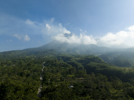 Aerial view of Mount Merapi volcano surrounded by lush green forests and clouds under a clear blue sky in Kecamatan Cangkringan Daerah Istimewa Yogya Indonesia. Aerial view of Mount Merapi volcano surrounded by lush green forests and clouds under a clear blue sky in Kecamatan Cangkringan Daerah Istimewa Yogya Indonesia.