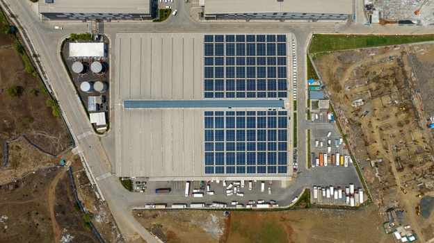 Aerial view of a large warehouse featuring a significant solar panel array on its roof surrounded by parking areas with trucks and industrial infrastructure in Bhiwandi Maharashtra India. Aerial view of a large warehouse featuring a significant solar panel array on its roof surrounded by parking areas with trucks and industrial infrastructure in Bhiwandi Maharashtra India.