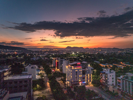 Aerial view of the cityscape of Udaipur at sunset featuring modern buildings lush greenery and distant mountains under a vibrant orange and purple sky in Udaipur Rajasthan India.