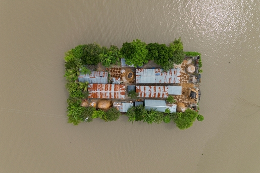 Mithamain Upazila Bangladesh 09 May 2026 Aerial view of a small isolated homestead island with corrugated metal roofs and lush green trees surrounded by floodwaters.