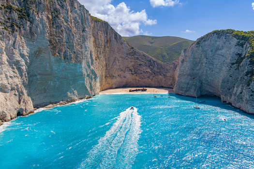 Aerial view of Navagio beach with its iconic shipwreck surrounded by towering limestone cliffs and turquoise waters with a boat leaving a white wake in Zakynthos Navagio Greece.
