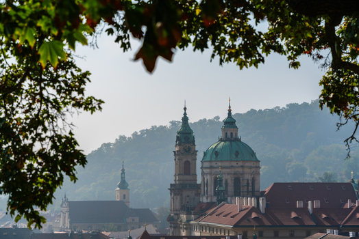 View of St. Nicholas Church with its green dome and bell tower framed by tree branches overlooking the historic rooftops of Prague Prague Czechia.