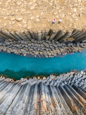 Aerial view of Studlagil Canyon with its iconic basalt columns and turquoise river with two people standing on the edge in Iceland. Aerial view of Studlagil Canyon with its iconic basalt columns and turquoise river with two people standing on the edge in Iceland.