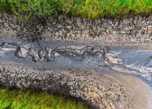Aerial view of a winding muddy stream bed with dark sediment patterns and green grass along the banks in Latvia.