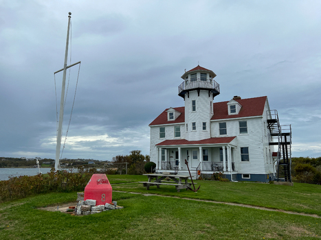 View of the historic Block Island Range Light with white wooden exterior and red roof set on a grassy lawn with a red buoy in the foreground in New Shoreham Rhode Island United States.