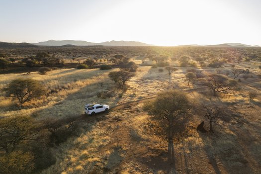 Aerial view of a white car navigates a dirt track through arid grasslands the setting sun casting long shadows across the Namibian landscape Windhoek Namibia. Aerial view of a white car navigates a dirt track through arid grasslands the setting sun casting long shadows across the Namibian landscape Windhoek Namibia.