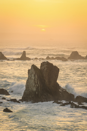 View of Silence Beach featuring jagged rock formations and crashing waves under a golden sunset sky in Asturias Spain.