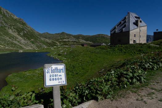 View of the Sankt Gotthard Pass featuring a historic stone building and a small alpine lake surrounded by green slopes and mountain peaks in Airolo Ticino Switzerland.