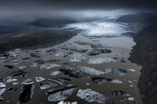 Aerial view of a massive glacier tongue flowing into a lagoon filled with floating icebergs under a dark moody sky in Iceland.