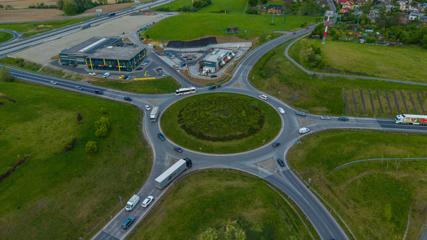 Aerial view of a busy traffic roundabout with cars and trucks moving through an intersection near commercial buildings and green fields. Aerial view of a busy traffic roundabout with cars and trucks moving through an intersection near commercial buildings and green fields.