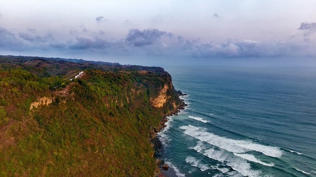 Aerial view of Giricahyo coastal cliffs with lush green vegetation meeting the ocean with rolling waves under a cloudy sky in Purwosari Special Region of Yogyakarta Indonesia. Aerial view of Giricahyo coastal cliffs with lush green vegetation meeting the ocean with rolling waves under a cloudy sky in Purwosari Special Region of Yogyakarta Indonesia.