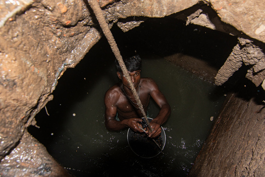 Dhaka Bangladesh 18 April 2018 View of a sewer cleaner working inside a dark manhole filled with murky water and mud holding a rope and bucket. Dhaka Bangladesh 18 April 2018 View of a sewer cleaner working inside a dark manhole filled with murky water and mud holding a rope and bucket.