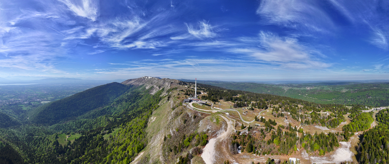 Aerial view of the Col de la Faucille mountain pass featuring a telecommunications tower winding roads and lush green forests under a blue sky with wispy clouds in Gex Auvergne Rhone Alpes France. Aerial view of the Col de la Faucille mountain pass featuring a telecommunications tower winding roads and lush green forests under a blue sky with wispy clouds in Gex Auvergne Rhone Alpes France.