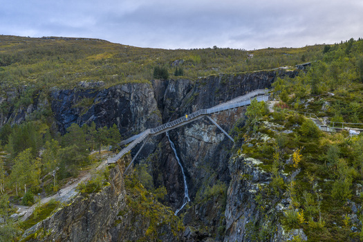 Aerial view of the Voringfossen waterfall and the modern pedestrian bridge spanning a deep rocky gorge with a person in a yellow jacket in Eidfjord Municipality Vestland Norway.
