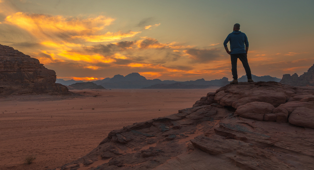 View of a man standing on a rocky outcrop overlooking the desert landscape of the Wadi Rum Protected Area at sunset with vibrant clouds Wadi Rum Village Aqaba Governorate Jordan. View of a man standing on a rocky outcrop overlooking the desert landscape of the Wadi Rum Protected Area at sunset with vibrant clouds Wadi Rum Village Aqaba Governorate Jordan.