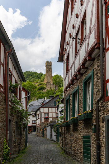 View of a narrow cobblestone street lined with traditional half timbered houses leading towards a medieval castle tower on a lush green hill under a cloudy sky in Monreal Rhineland Palatinate Germany. View of a narrow cobblestone street lined with traditional half timbered houses leading towards a medieval castle tower on a lush green hill under a cloudy sky in Monreal Rhineland Palatinate Germany.
