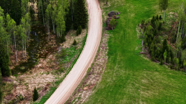 Aerial view of a winding dirt road cutting through a lush green forest and open meadows in Latvia. Aerial view of a winding dirt road cutting through a lush green forest and open meadows in Latvia.