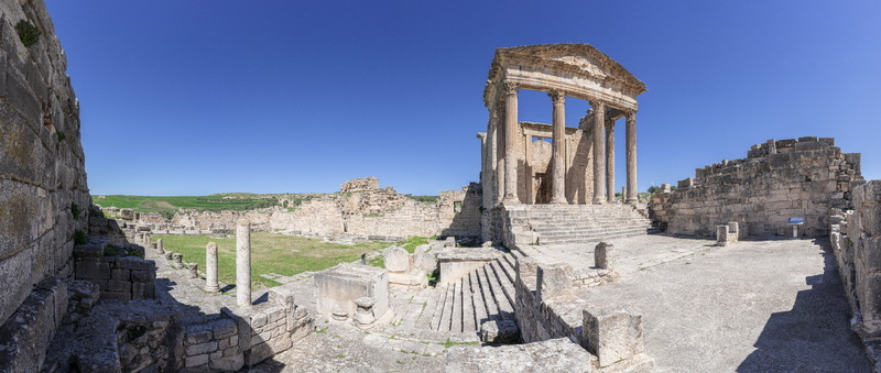 View of the Capitol temple at the Dougga Archaeological Site with ancient stone columns ruins and green fields under a clear blue sky in Teboursouk Beja Governorate Tunisia.