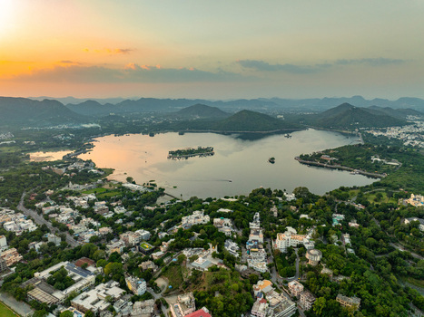 Aerial view of Lake Pichola with the island palace surrounded by the cityscape lush greenery and distant hills under a warm sunset sky in Udaipur Rajasthan India.