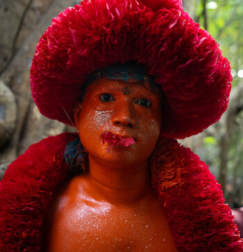 Munshiganj Bangladesh 13 April 2025 View of a person with vibrant red body paint and a fluffy headdress their face highlighted amidst blurred green foliage. Munshiganj Bangladesh 13 April 2025 View of a person with vibrant red body paint and a fluffy headdress their face highlighted amidst blurred green foliage.