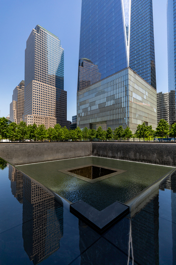 View of the 911 Memorial reflecting pool with One World Trade Center rising in the background surrounded by trees and modern skyscrapers in New York New York United States. View of the 911 Memorial reflecting pool with One World Trade Center rising in the background surrounded by trees and modern skyscrapers in New York New York United States.
