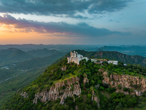 Aerial view of the historic Sajjangarh Palace perched atop a lush green hill in the Aravalli range during a vibrant sunset in Udaipur Rajasthan India.