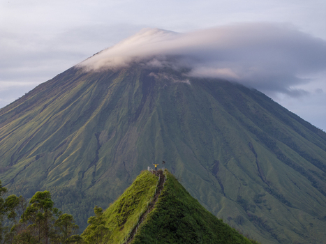 Aerial view of Mount Inerie with a person standing on a lush green ridge dramatic volcanic slopes and a cloud cap in Inerie Flores Indonesia. Aerial view of Mount Inerie with a person standing on a lush green ridge dramatic volcanic slopes and a cloud cap in Inerie Flores Indonesia.