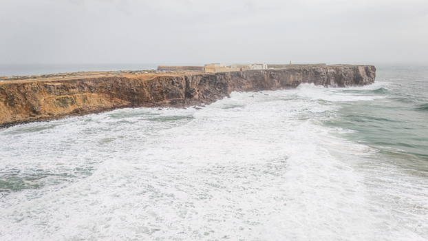 Aerial view of the historic Sagres Fortress perched on high rocky cliffs with powerful Atlantic waves crashing against the shore under an overcast sky in Sagres Faro Portugal. Aerial view of the historic Sagres Fortress perched on high rocky cliffs with powerful Atlantic waves crashing against the shore under an overcast sky in Sagres Faro Portugal.