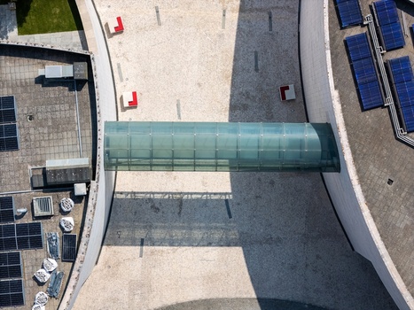 Aerial view of a modern building featuring a glass bridge connecting two curved sections with rooftop solar panels and a paved courtyard in Lisboa Lisboa Portugal.