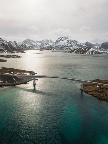 Aerial view of a sleek bridge elegantly spans the turquoise waters contrasting with the rugged snow dusted mountains in the distance Flakstadbruene Norway. Aerial view of a sleek bridge elegantly spans the turquoise waters contrasting with the rugged snow dusted mountains in the distance Flakstadbruene Norway.