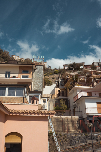 View of buildings cascading down the hillside under a dramatic sky leading up to the illuminated Hotel sign Taormina Sicily Italy.