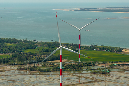 Aerial view of towering wind turbines standing tall against the backdrop of the calm sea with green fields and lush trees Cox s Bazar Chittagong Division Bangladesh. Aerial view of towering wind turbines standing tall against the backdrop of the calm sea with green fields and lush trees Cox s Bazar Chittagong Division Bangladesh.