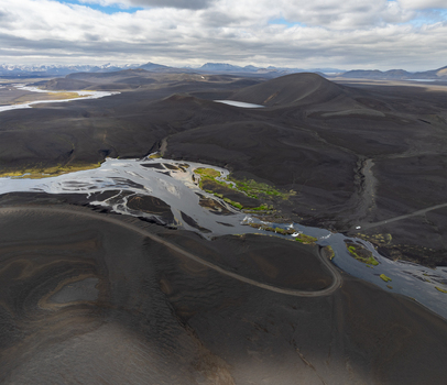 Aerial view of a braided river flowing through a vast volcanic landscape with black sand dark hills and patches of green moss under a cloudy sky in Iceland. Aerial view of a braided river flowing through a vast volcanic landscape with black sand dark hills and patches of green moss under a cloudy sky in Iceland.