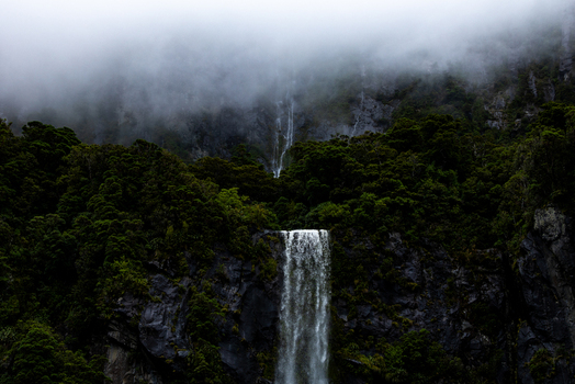 View of a dramatic waterfall cascading down a dark rocky cliff face shrouded in mist and lush vegetation creating an ethereal atmosphere Milford Sound New Zealand.