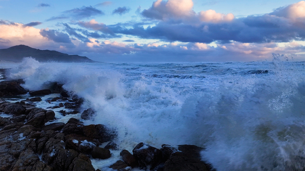 Aerial view of rough ocean waves crashing against a rocky shoreline under a dramatic cloudy sky with distant mountains Cape Town Western Cape South Africa. Aerial view of rough ocean waves crashing against a rocky shoreline under a dramatic cloudy sky with distant mountains Cape Town Western Cape South Africa.