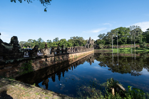 View of the historic stone causeway and moat leading to the Angkor Wat temple complex surrounded by lush trees under a clear blue sky in Krong Siem Reap Siem Reap Province Cambodia. View of the historic stone causeway and moat leading to the Angkor Wat temple complex surrounded by lush trees under a clear blue sky in Krong Siem Reap Siem Reap Province Cambodia.