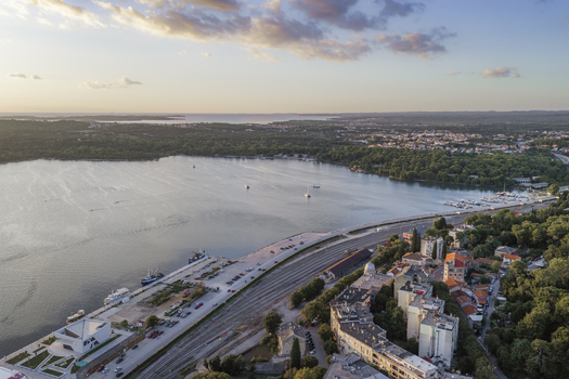 Aerial view of the coastal harbor with boats railway tracks urban buildings and lush greenery under a soft sunset sky in Pula Istarska zupanija Croatia. Aerial view of the coastal harbor with boats railway tracks urban buildings and lush greenery under a soft sunset sky in Pula Istarska zupanija Croatia.