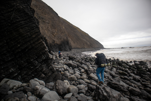 View of hikers with backpacks walking along the rocky shoreline of the Lost Coast beneath dark layered cliffs in Humboldt County California United States.