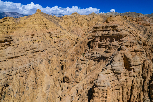 Aerial view of rugged golden brown mountain rock formations with deep canyons and layered sedimentary cliffs under a clear blue sky in Nepal.
