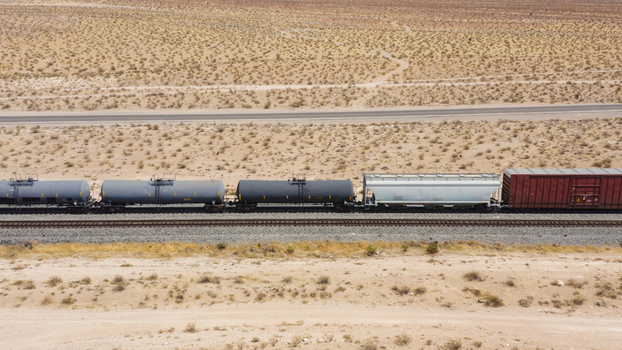 Aerial view of a freight train with various railcars including tank cars and a boxcar traveling through a vast desert landscape in the United States.