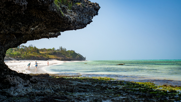 View of clear turquoise waters gently lap against the white sandy beach shadowed by rugged rock formations inviting exploration in Paje Unguja South Region Tanzania.