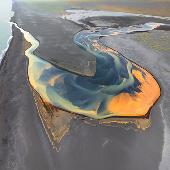 Aerial view of a glacial river delta with vibrant blue and orange sediment patterns flowing into the sea across black volcanic sand in Iceland. Aerial view of a glacial river delta with vibrant blue and orange sediment patterns flowing into the sea across black volcanic sand in Iceland.