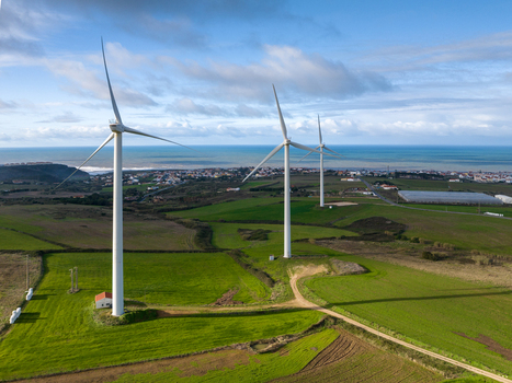 Aerial view of three wind turbines standing in green fields overlooking a coastal town and the Atlantic Ocean under a cloudy sky Ribamar Portugal. Aerial view of three wind turbines standing in green fields overlooking a coastal town and the Atlantic Ocean under a cloudy sky Ribamar Portugal.