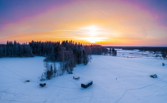 Aerial view of a snowy landscape at sunset featuring a small wooden structure a group of people standing nearby and a dense forest under a vibrant sky in Latvia. Aerial view of a snowy landscape at sunset featuring a small wooden structure a group of people standing nearby and a dense forest under a vibrant sky in Latvia.
