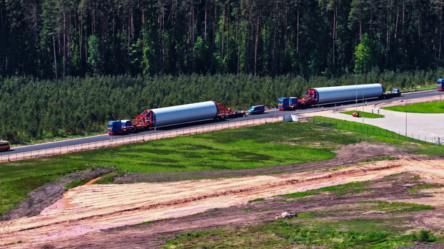Aerial view of heavy transport trucks carrying large wind turbine tower sections along a road bordered by a dense pine forest in Latvija. Aerial view of heavy transport trucks carrying large wind turbine tower sections along a road bordered by a dense pine forest in Latvija.
