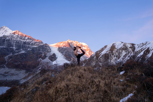 Aerial view of a man performing a yoga dancer pose on a grassy ridge with snow capped Himalayan peaks illuminated by sunrise in Nepal.