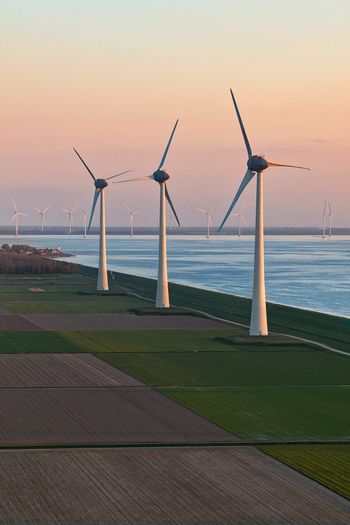 Aerial view of wind turbines standing along a coastal dike next to agricultural fields under a soft sunset sky Netherlands.
